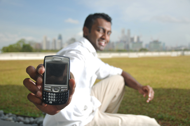 IMAGE: Indian Man dressed in white shirt and khaki pants smiles as he holds out his feature phone.
